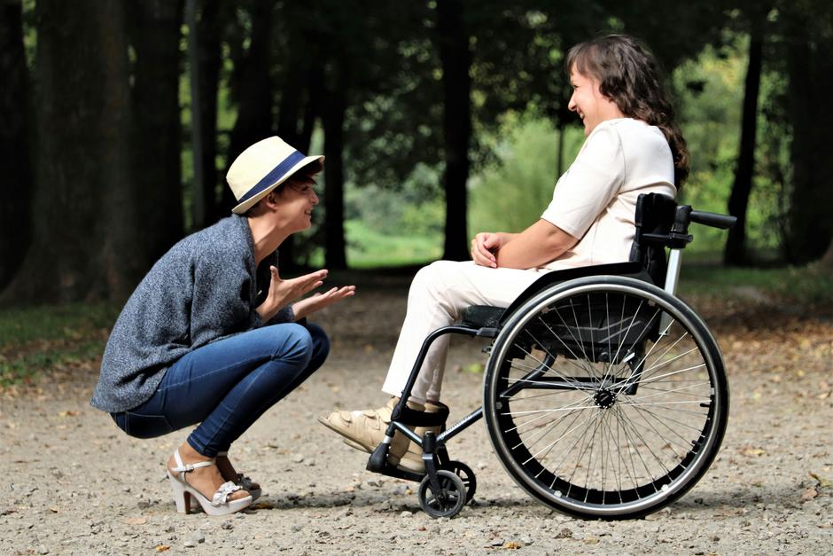 Osoba na wózku inwalidzkim uśmiecha się, spacerując po parku z przyjacielem.